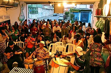 Grupo de pessoas reunido em um espaço cultural, com músicos tocando instrumentos de percussão e cordas.