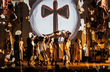 Apresentação teatral com grupo de atores em trajes tradicionais e fundo projetado com símbolo de uma cruz estilizada.