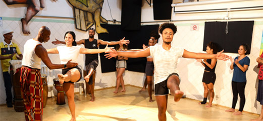 Grupo de pessoas participando de uma oficina de dança, com orientador guiando os movimentos em um espaço cultural.