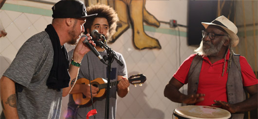 Trio musical durante uma apresentação, com um cantor, um músico tocando cavaquinho e outro tocando tambor, em um espaço cultural.