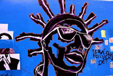 Mural artístico com retrato estilizado de uma figura com cabelo de dread e óculos, sobre fundo azul com colagens e grafites.