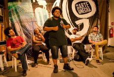 Participantes de um debate no AGÔ, com uma pessoa falando no microfone e outros sentados em uma mesa, em frente a um fundo colorido.
