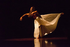 Dançarina em movimento fluido e expressivo no palco, com saia longa e iluminada contra um fundo escuro.