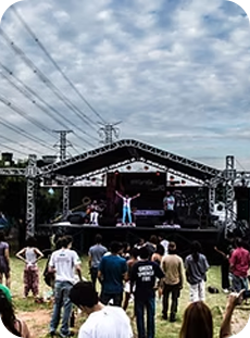 Apresentação musical ao ar livre em um palco montado com estrutura metálica, com público assistindo em um gramado sob um céu parcialmente nublado.