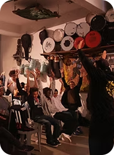 Grupo de pessoas em um espaço cultural com instrumentos pendurados nas paredes, participando de uma atividade coletiva com braços levantados.