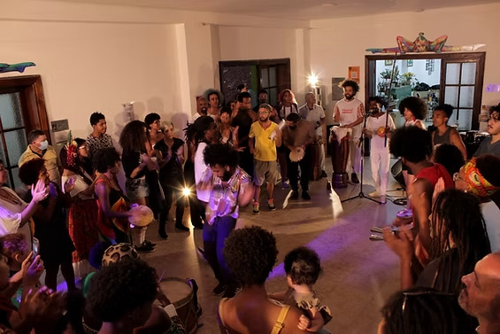 Grupo de pessoas reunidas em um espaço interno participando de uma roda de música e dança com instrumentos de percussão.