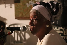 Mulher idosa com turbante, falando ao microfone, em uma apresentação no Encontro de Griôs.