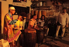 Grupo de músicos se apresentando em palco íntimo, com destaque para instrumentos de percussão e um cantor usando vestes coloridas.