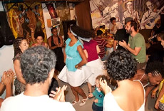 Pessoas dançando animadamente em um evento da Escola de Samba Totonho, com destaque para uma mulher girando sua saia e a interação alegre do público.