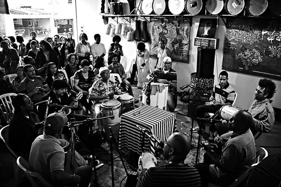Grupo de músicos tocando instrumentos de percussão enquanto o público, ao fundo, assiste atento a uma apresentação ao vivo.
