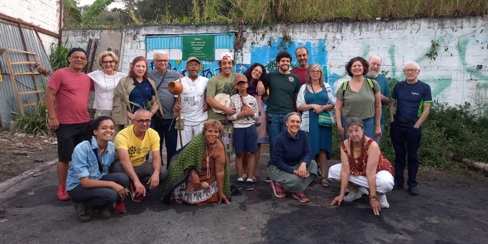 Grupo de pessoas sorrindo e posando para foto ao ar livre, com instrumentos de percussão, em frente a um muro grafitado.
