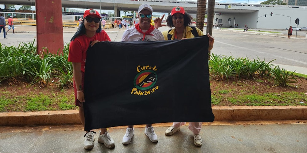 Três membros do Círculo Palmarino segurando uma bandeira do grupo, sorrindo e em trajes característicos, com o logotipo do Círculo Palmarino visível.