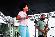 Cantora se apresentando no palco com microfone, acompanhada de músico tocando saxofone.