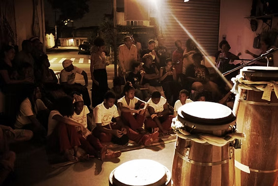 Público assistindo a uma apresentação musical em um sarau, com tambores de percussão em primeiro plano e iluminação suave.