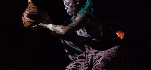 Performer segurando um instrumento de cabaça durante uma apresentação artística com iluminação dramática.